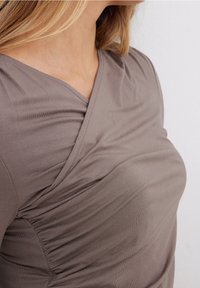 Primo piano di una donna che indossa un top avvolgente color taupe con ruches e scollatura a V, che mostra la spalla e la parte superiore del petto.