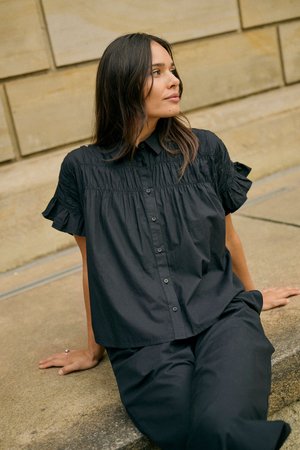 Femme aux cheveux bruns portant un chemisier noir boutonné et un pantalon, assise sur des marches en pierre, regardant sur le côté.