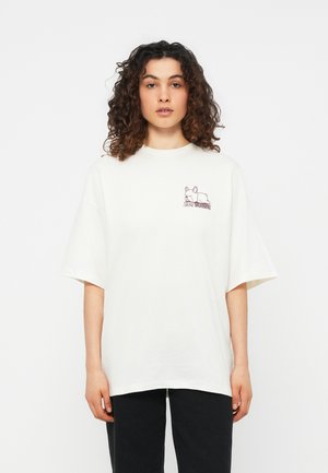 Γυναίκα με σγουρά μαλλιά φοράει υπερμεγέθη λευκό μπλουζάκι με μικρό κοιμισμένο σκύλο και τη φράση «Slow Morning», συνδυασμένο με μαύρο παντελόνι.