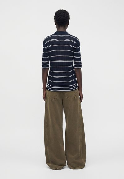 Personne de dos portant une chemise rayée bleu marine et blanc à manches mi-longues et un pantalon large vert olive.