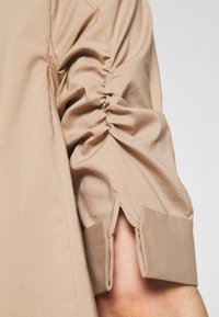 Manica di camicia beige con dettaglio arricciato e apertura al polsino. Tessuto liscio con una consistenza morbida e una vestibilità sartoriale. Design semplice senza motivi.