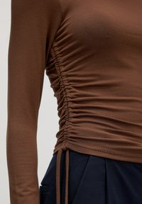 Bruine top met lange mouwen, voorzien van een geplooid zijdetail en een verstelbare trekkoord. De stof heeft een gladde textuur en een getailleerd ontwerp.