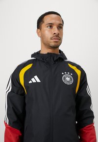 Mand med sort, rød og gul sportsjakke med lynlåskrave, Adidas-logo og tysk fodboldforeningsmærke på brystet.