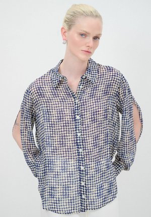 Camicia trasparente con bottoni di colore crema chiaro, decorata con un motivo a pois blu. Presenta un colletto classico e maniche ampie con fessure laterali.