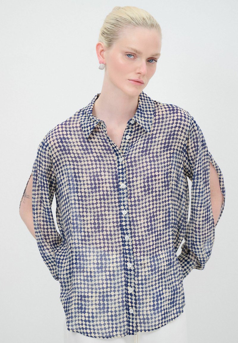 Camicia trasparente con bottoni di colore crema chiaro, decorata con un motivo a pois blu. Presenta un colletto classico e maniche ampie con fessure laterali.