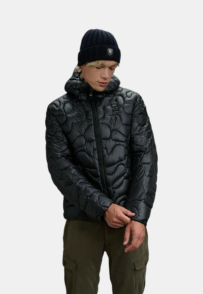 Giacca a vento nera con motivo trapuntato, chiusura con zip e cappuccio. Indossata con un berretto a coste scuro, abbinata a pantaloni cargo verde oliva.