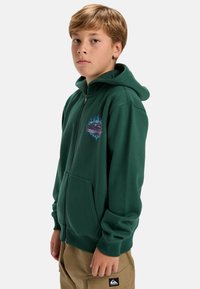 Sudadera verde con cremallera, hecha de un material suave, con un bolsillo frontal y un diseño gráfico en el pecho, combinada con pantalones beige.
