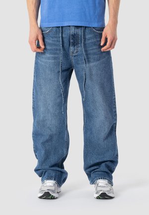 Uomo che indossa jeans di denim azzurro larghi con vita a coulisse e sneakers bianche, in piedi contro uno sfondo neutro.