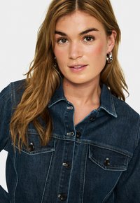 Frau mit hellbraunem Haar, die silberne Creolen und eine dunkelblaue Jeansjacke mit schwarzen Knöpfen trägt, vor einem einfarbigen Hintergrund.