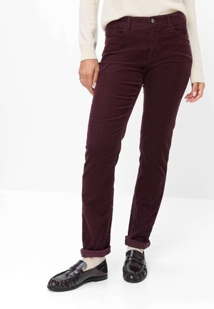 Person iført opkørte mørkerøde bukser, cremfargede sokker, sorte loafers og en cremefarvet langærmet top, stående mod en hvid baggrund.