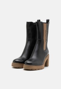 Bottes chelsea en cuir noir avec des talons épais couleur tan, comportant un panneau latéral marqué "GMK." Texture lisse et design élégant.