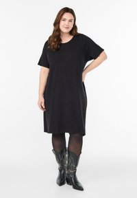 Musta lyhythihainen mekko pehmeästä kankaasta, ylettyen kesäreiteen. Päälle on puettu mustat teksturoidut sukkahousut ja koristellut cowboy-saappaat.