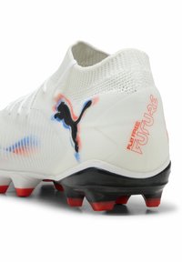 Botas de fútbol blancas con parte superior de tejido texturizado, logo de Puma negro, acentos rojos y azules, y suela negra con tacos rojos.
