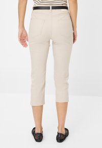 Beige cropped broek met een gladde textuur en slim fit, voorzien van twee achterzakken en afgewerkte manchetten, gecombineerd met zwarte mesh schoenen.