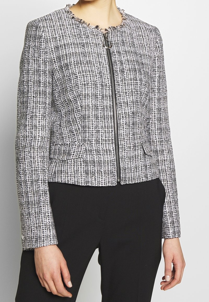 Femme portant une veste en tweed texturée noir et blanc avec fermeture éclair devant et un pantalon noir sur mesure sur un fond blanc uni.