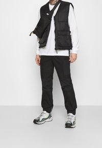 Schwarze gepolsterte Weste mit mehreren Taschen, getragen über einem weißen Langarmshirt. Schwarze Cargo-Hosen und graue Turnschuhe mit grünen Akzenten runden den Look ab.