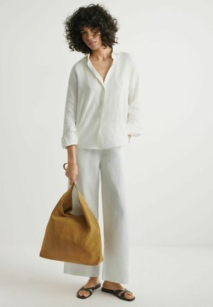 Kobieta o kręconych włosach, ubrana w luźną białą lnianą koszulę i spodnie, trzymająca dużą brązową zamszową torbę, stojąca na białym tle.