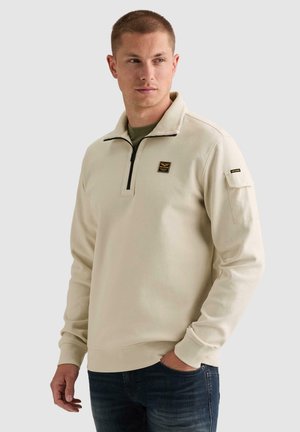 Man wearing beige quarter-zip sweatshirt with small patches on chest and sleeve, dark jeans, standing against a plain light background.