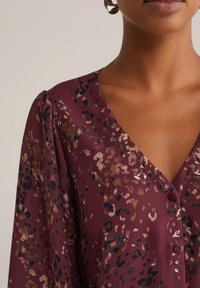 Blouse bordeaux avec un motif léopard en or, bronze et noir. Dotée d'un décolleté en V et de manches légèrement bouffantes avec des boutons sur le devant.