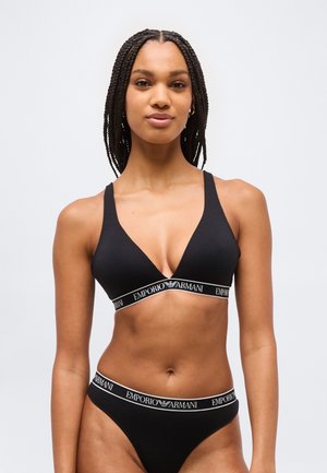 Femme aux cheveux tressés portant un soutien-gorge noir de la marque Emporio Armani et une culotte assortie, debout devant un fond clair uni.
