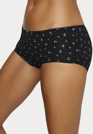 Frau trägt schwarze Boyshort-Unterwäsche mit kleinem blauem Blumenmuster und weißen Punkten, steht vor einem einfarbigen Hintergrund.