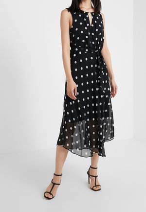 Vrouw draagt een mouwloze zwarte midi-jurk met witte polka dots, een taillekoord en zwarte sandalen met hoge hakken, staand op een witte vloer.