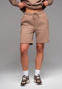 Pantaloni corti beige in cotone con vita elasticizzata e cordino, dotati di tasche laterali. Abbinate a sneaker con motivo e calze bianche alla caviglia.
