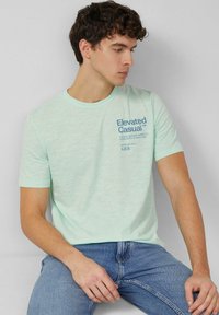 Licht muntgroen t-shirt met korte mouwen, gemaakt van zacht stof, met een blauwe bedrukte tekst op de voorkant. Gecombineerd met blauwe denim jeans.