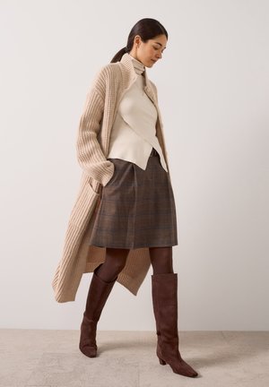 Femme portant un long cardigan beige en maille, un haut à col roulé crème, une jupe à carreaux marron, des collants noirs et des bottes marron montant jusqu'aux genoux, marchant à l'intérieur.