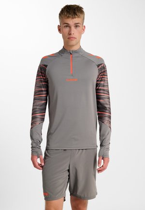 Jeune homme portant un haut de sport gris avec manches à motifs et un short assorti, debout devant un fond blanc uni.