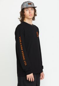 Schwarzes Langarmshirt mit orangefarbener Markenlogo auf der Brust und am Ärmel, in einem lässigen Schnitt. Das Modell trägt eine gemusterte graue Mütze.