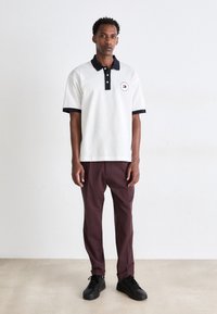 Polo blanc avec un col et une bordure bleu marine, arborant un logo circulaire. Assorti à un pantalon bordeaux et des chaussures noires, posé sur une surface neutre.