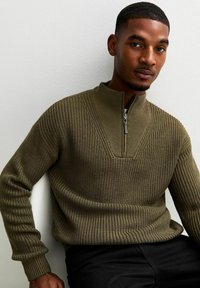 Olivgrön stickad tröja med halvdragkedja, med en v-ringad design och ribbad textur. Buren med svarta byxor, sittande mot en vägg.