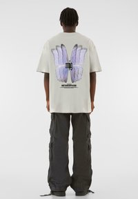 Lys grå oversized t-shirt med et stort lilla sommerfuglegrafik på ryggen, parret med mørke cargo-bukser med sidelommer.