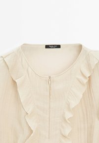 Blouse beige clair avec col rond, fermeture éclair centrale et détails à volants sur le devant et les épaules.