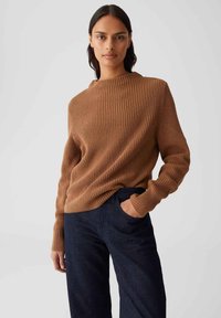 Frau in einem braunen gerippten Pullover und dunklen blauen Jeans, mit einer Hand in der Tasche, steht vor einem einfarbigen hellen Hintergrund.