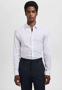 Camisa blanca de manga larga con cuello clásico, frente abotonado y textura suave, combinada con pantalones de color oscuro.