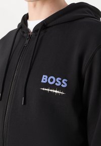 Felpa nera con zip realizzata in tessuto morbido, con logo blu "BOSS" e grafica di un'onda sonora bianca sul lato sinistro del petto.