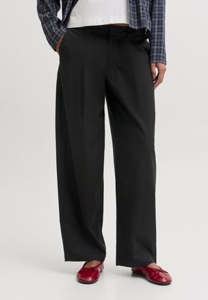 Personne portant un pantalon noir à jambes larges, des ballerines rouges, un haut blanc et une veste à carreaux bleue, debout les mains dans les poches.