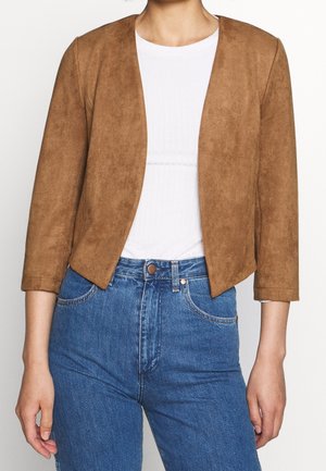 Femme portant une veste courte en daim marron ouverte devant avec des manches trois-quarts sur un haut blanc et un jean bleu.