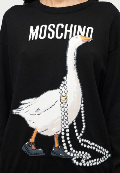 Μαύρο πλεκτό πουλόβερ με γραφικό λευκού χήνου που φοράει μαύρα παπούτσια και περιδέραιο με πέρλες. Το λογότυπο «Moschino» εμφανίζεται πάνω από το σχέδιο.