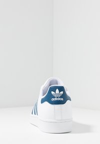 White sneaker with blue accents, featuring three side stripes, a textured heel tab, and a rubber sole. Logo displayed on the heel.