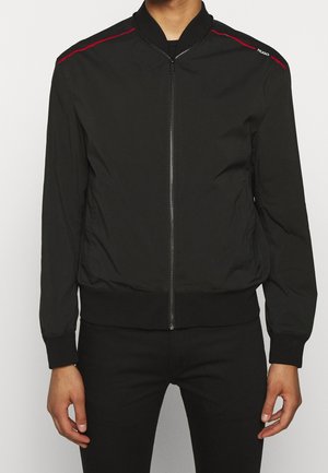 Czarna kurtka bomberka na zamek z prążkowanym kołnierzem i mankietami, czerwonymi akcentami na ramionach oraz logo "Hugo" na prawym ramieniu, noszona przez stojącą osobę.