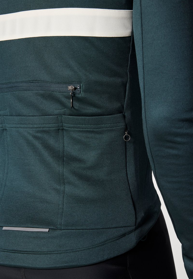 Dark green athletic jacket with a white horizontal stripe, featuring a zippered pocket and textured fabric. Long sleeves and curved hem design.