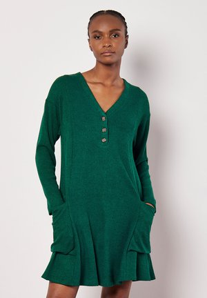 Grünes geripptes Kleid mit V-Ausschnitt, langen Ärmeln, einem drei-knöpfigen Streifen und seitlichen Taschen, mit einem ausgestellten Saum und weicher Textur.