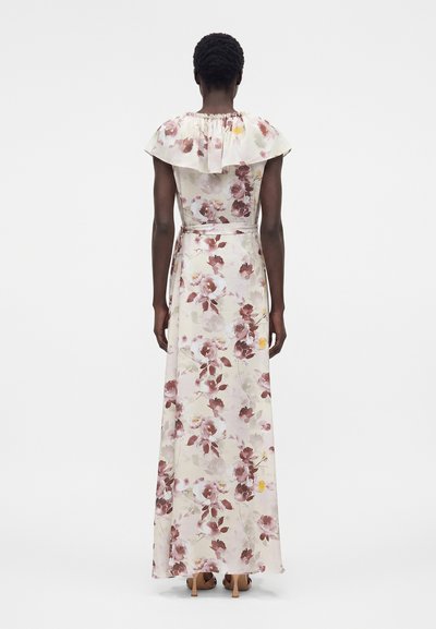 Γυναίκα που φοράει μακρύ, αμάνικο, φλοράλ φόρεμα με βολάν στον γιακά, στέκεται γυρισμένη με την πλάτη σε απλό λευκό φόντο.