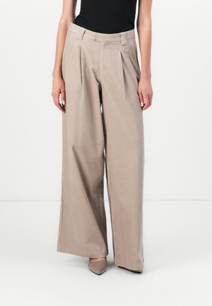 Beige weit geschnittene Hose mit vorderen Falten und Gürtelschlaufen, kombiniert mit einem schwarzen Oberteil und spitz zulaufenden Schuhen vor weißem Hintergrund.