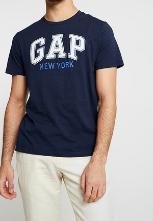 Homme portant un t-shirt GAP New York bleu marine et un pantalon blanc cassé, debout avec le bras gauche détendu et la main droite légèrement ouverte.