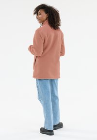 Fleecejacke in sanftem Korallenpink, mit hohem Kragen und seitlichen Taschen, kombiniert mit hellblauen Jeans und schwarzem Schuhwerk.