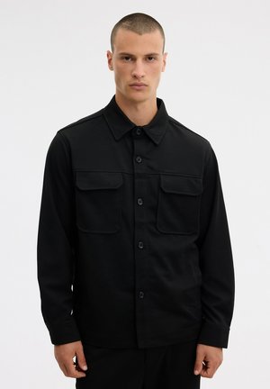 Un hombre luce una chaqueta camisera negra con botones delanteros y dos bolsillos de pecho con solapa sobre un fondo blanco liso.